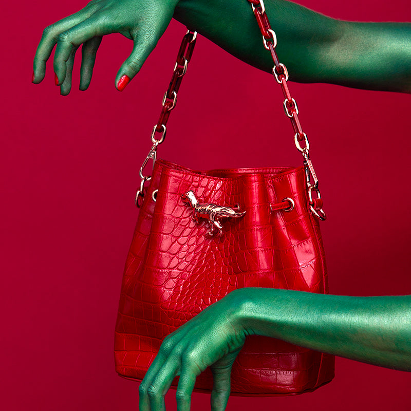 Jewel clip in the shape of a golden T-rex dinosaur on a red leather seal bag with green paint hands